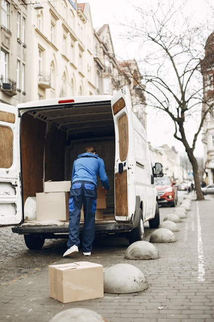 A man wearing a white polo shirt and black cap is standing outside next to a white moving van during daylight. He is holding a cardboard box and smiling at the camera. The van's rear doors are open, r