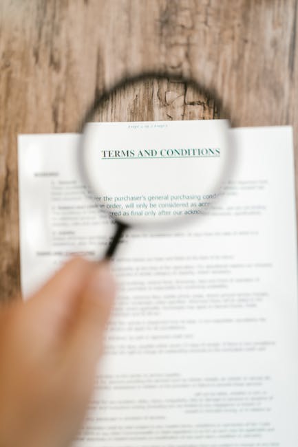A close-up image of a person holding a magnifying glass over a printed document titled 'Terms and Conditions,' with the magnifying glass focusing on the heading. The document is placed on a wooden surface, possibly a table or floor, and the person's hand is visible holding the handle of the magnifying glass. In the background, the blurred text of the rest of the document can be seen beneath the magnification. This scene suggests review or examination of paperwork related to a home relocation or removal service, with the magnifying glass emphasizing careful reading of contractual details. The setting and context imply an environment where detailed planning or agreement review occurs before the moving process, aligning with the services offered by companies like Man with Van Upper Clapton for furniture transport and packing during a house move.