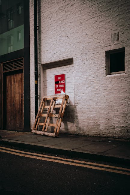 A wooden pallet leaning against a white brick wall on a sidewalk outside a building, with a red and white sign attached to the wall indicating 'No King in this Area'. The pallet appears unused and is positioned near a dark wooden door and a small rectangular window. The scene is illuminated by natural light, and the pavement shows double yellow lines along the curb, suggesting an urban street environment. This setting may be part of preparations for a home relocation or furniture transport process, with the pallet potentially used for moving or storing items during a removal service, as provided by companies like Man with Van Upper Clapton.