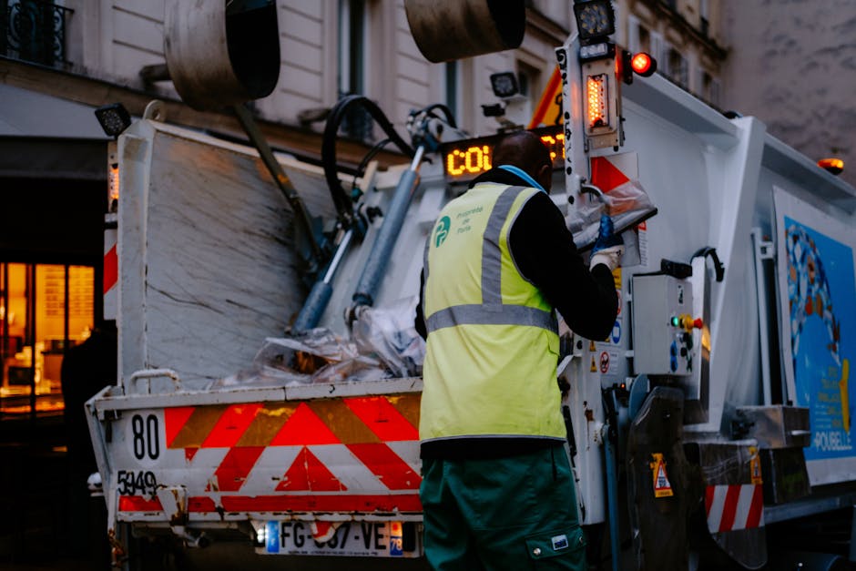A waste collection worker wearing a high-visibility yellow vest with reflective strips, dark trousers, and gloves is operating a mechanical compactor on a white refuse truck with orange and red reflective markings outdoors during evening or dusk. The worker is standing at the side of the truck, using controls to operate the compactor's mechanism for loading waste materials. The truck, branded with the company logo, is positioned on a pavement or driveway, and the background includes a building with illuminated windows. This scene illustrates waste collection and disposal processes relevant to home removal or clearance services, supported by Man with Van Upper Clapton’s expertise in trash or bulk waste removal in the Upper Clapton area.