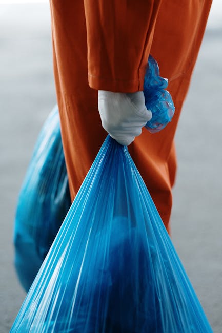 A person wearing an orange protective suit and white gloves is holding a large blue plastic garbage bag filled with waste, standing outdoors on a paved surface. In the background, there are additional similar bags, indicating waste collection or clearance activity, which is part of home relocation or clearance services. The scene suggests a cleanup or removal process, possibly associated with furniture, packaging materials, or household debris prepared for transport or disposal. This visual is relevant to house removals and packing services offered by Man with Van Upper Clapton, as part of their furniture transport, loading, or general clearance operations during a household move or waste removal project.
