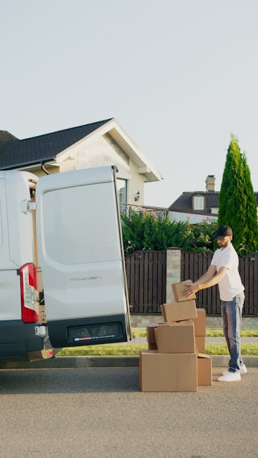 A man seated on a wooden chair outside a white delivery van parked on a city street during daytime, with the back doors open. The van's interior is lined with brown wooden panels and contains various