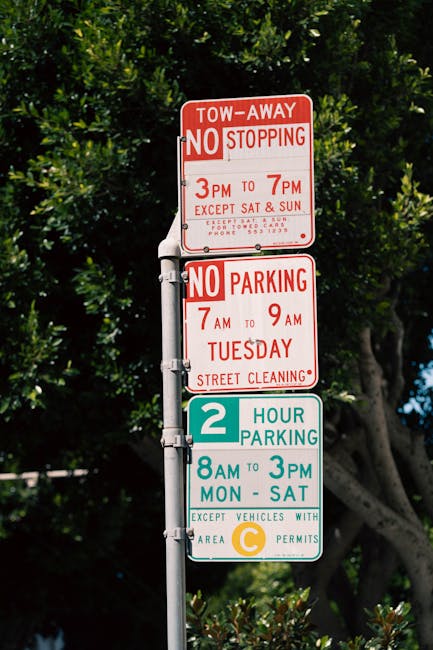 A vertical metal street signpost displaying multiple parking and stopping restrictions in Hackney. The top sign indicates a tow-away zone with no stopping from 3 pm to 7 pm, except Saturdays and Sundays, with additional instructions for towing and contact details. Below, a second sign prohibits parking from 7 am to 9 am on Tuesdays for street cleaning. The third sign shows a two-hour parking limit from 8 am to 3 pm, Monday to Saturday, for vehicles with an area C parking permit. The signs are affixed to a pole surrounded by lush green trees and foliage, with a glimpse of blue sky in the background, situated in an urban setting typical of Upper Clapton. The signpost is relevant to removals and moving services by informing about parking regulations that may affect home relocation or furniture transport in the area, as managed by Man with Van Upper Clapton.
