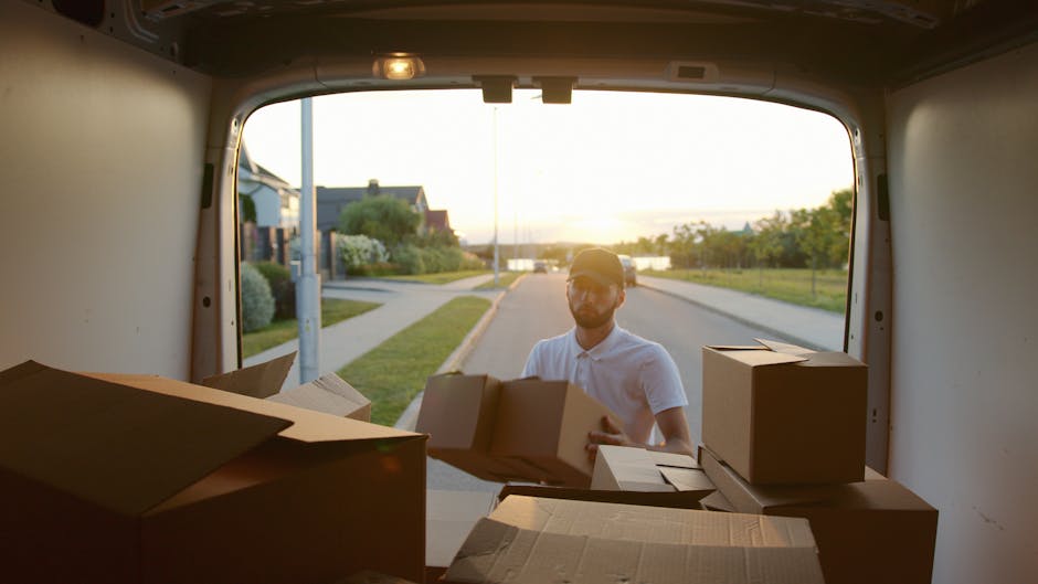 A man wearing glasses and a light-colored polo shirt is standing outside a white moving van, loading or unloading cardboard boxes. The photo is taken from inside the van, showing the open back door and the interior space filled with packed cardboard boxes of various sizes, some with tape, stacked near the edge of the vehicle. Outside, in the background, there is a residential street with houses, a sidewalk, and a lamp post, illuminated by warm, late-afternoon sunlight. The scene depicts a home relocation or furniture transport process, with the man engaged in packing or unpacking as part of a professional removals service, such as that provided by Man with Van Upper Clapton, supporting efficient and organised house move logistics in the Upper Clapton area. The environment appears calm and suburban, with greenery and a distant body of water visible on the horizon, suggesting a peaceful neighborhood setting.