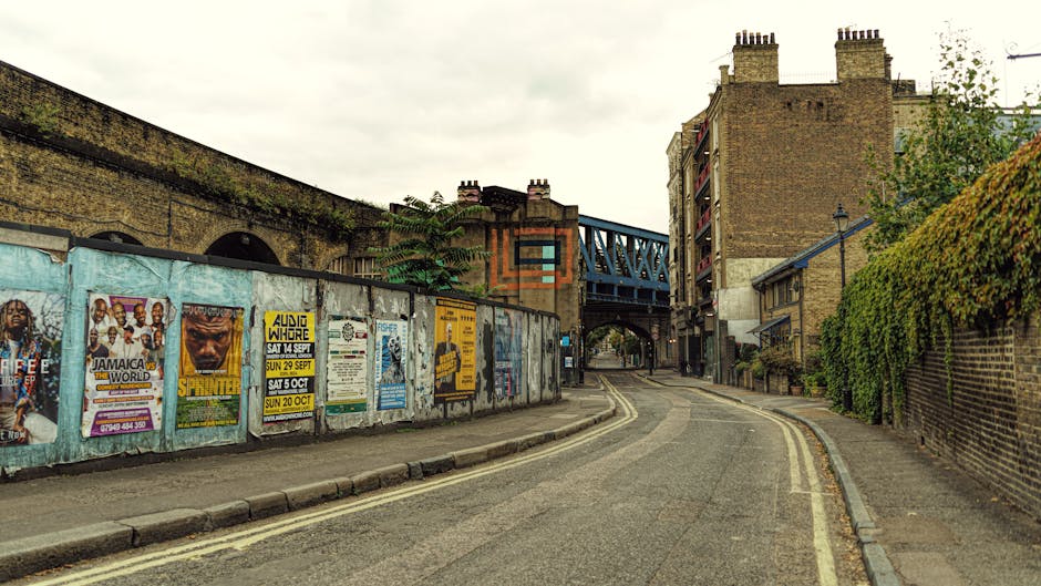 A narrow residential street with a slight curve in Upper Clapton, showing brick buildings on the right side partially covered by green ivy and trees, and a concrete wall on the left side covered with colorful posters advertising local events and music gigs. The pavement is lined with a curb, and a double yellow line runs along the edge of the road, indicating parking restrictions. An elevated blue metal bridge structure crosses over the street in the background, with a building featuring multiple chimneys visible behind it. The sky is overcast, casting diffuse natural light across the scene. This setting reflects an urban environment suitable for house removals and furniture transport, with street-level access for loading or unloading in a typical home relocation process, as performed by companies like Man with Van Upper Clapton.