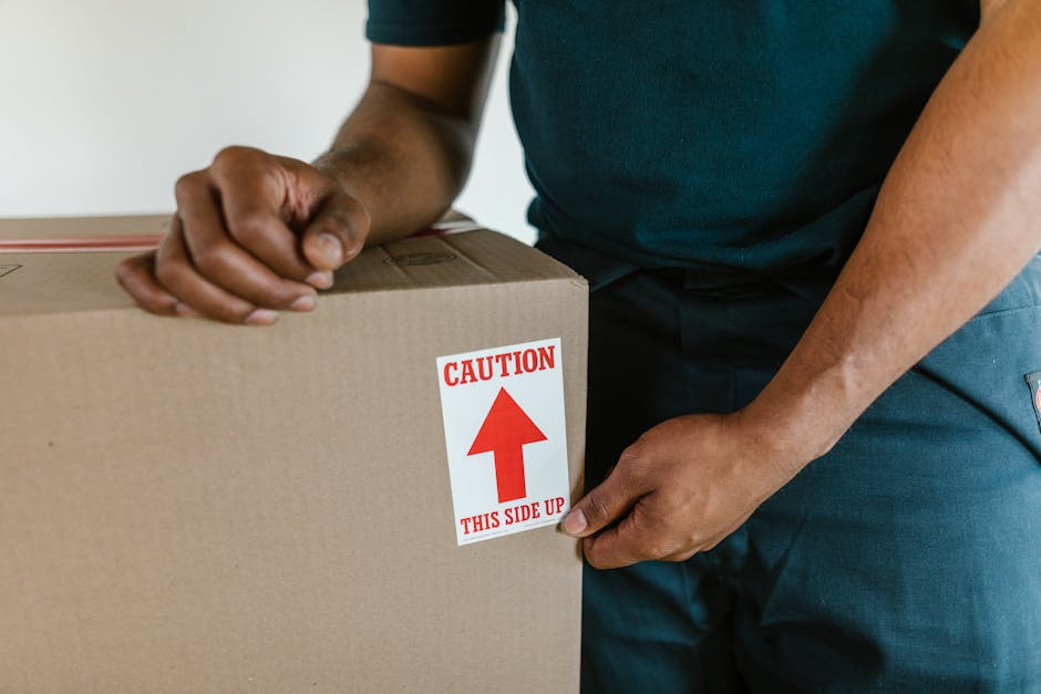 Close-up of a person wearing a dark blue shirt and matching pants, holding a large cardboard box with both hands. The box has a red and white caution label indicating 'THIS SIDE UP' with an upward-pointing arrow. The person is indoors near a doorway or entrance area, which is part of a home or flat undergoing packing or moving preparations. The scene suggests careful handling of household items during a house removal or relocation process facilitated by Man with Van Upper Clapton, consistent with the services offered in their UPPER CLAPTON removals guide. The background shows a neutral-colored wall and a glimpse of the floor, emphasizing the focus on the packing activity and the importance of correct handling during furniture transport and home relocation tasks.