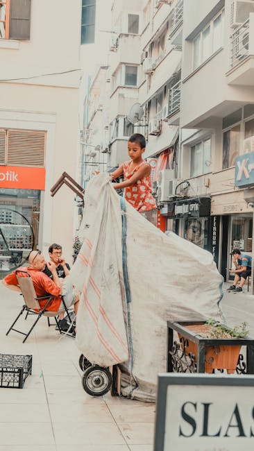 A young boy with short dark hair, wearing a sleeveless red top with white patterns, stands on a small platform or trolley with large gray fabric-covered sides, lifting or adjusting a large white and orange striped fabric cover. The scene is set on a busy urban sidewalk in a high-density residential and commercial area, with multi-storey buildings featuring balconies and windows overhead. Several pedestrians are seated on chairs along the pavement, some taking photos or observing the activity, while others walk past on the street. A potted plant in an orange container is visible in the foreground, and storefront signage, including a partially visible sign reading 'SLASH', indicates nearby businesses. The environment appears brightly lit with natural daylight, and the overall context suggests a home relocation or furniture transport scenario involving careful handling and use of protective coverings during the moving process. The setting reflects the logistical aspect of street-based furniture or appliance moving services provided by Man with Van Upper Clapton, emphasizing the challenges of navigating narrow streets and urban environments during house removals.