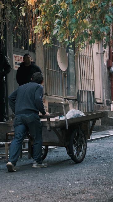 Two men are engaged in a home relocation process on a narrow street in Clapton, with one man pushing a large, black, three-wheeled self-propelled trolley loaded with wrapped furniture and cardboard boxes, while the other man, standing nearby, appears to coordinate or supervise. The trolley is positioned close to a building entrance with a metal gate and a satellite dish attached to the wall. The scene takes place outdoors on a paved street with some greenery overhead, casting shadows, indicating it is daytime. The men are dressed casually, and the presence of packing materials suggests an organized furniture transport or moving service by Man with Van Upper Clapton, part of professional removals. Items are carefully wrapped in fabric or plastic to protect during loading and transport, illustrating the logistical aspects involved in house removals through narrow urban streets, with careful handling of large household items during the loading process.