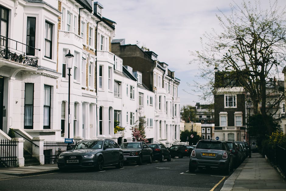 The image shows a residential street in Upper Clapton with white multi-storey terraced houses featuring bay windows, black railings, and front doors. Several cars, including an Audi and a Mini, are parked along the curbside on both sides of the road, which is lined with asphalt pavement. A tall, black street lamp is positioned near the front of the houses, and a large tree with sparse green foliage extends its branches over part of the street. Overcast skies cast diffuse light on the scene, providing visibility of the exterior details of the houses and parked vehicles. The context of the image relates to house removals and furniture transport, where loading and parking considerations near Clapton Pond are relevant, as illustrated by the parked vehicles ready for a potential relocation process, consistent with the services offered by Man with Van Upper Clapton.