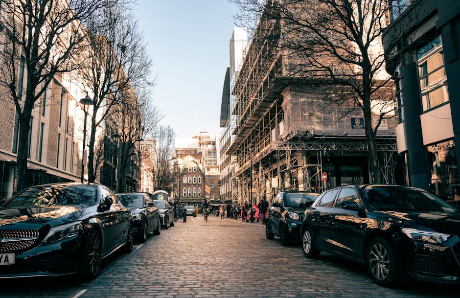 A street scene near Clapton Pond during daytime showing parked cars along the cobblestone pavement, including a black luxury sedan in the foreground, with additional vehicles lining both sides of the road. On the right, there is a building under construction with scaffolding and protective sheeting, while the left side features modern office or residential buildings with leafless trees lining the street. Pedestrians are visible walking in the distance, likely engaged in activities such as a home relocation or furniture transport. The lighting suggests late afternoon with natural sunlight casting shadows on the street and vehicles. The setting captures a typical urban environment where professional removal services, like those offered by Man with Van Upper Clapton, operate, potentially involving loading or unloading furniture or boxes from vehicles into nearby properties.