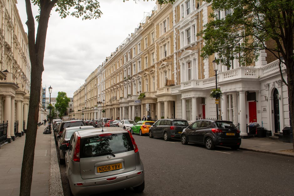 The image shows a residential street in Upper Clapton with white multi-storey terraced houses featuring bay windows, black railings, and front doors. Several cars, including an Audi and a Mini, are parked along the curbside on both sides of the road, which is lined with asphalt pavement. A tall, black street lamp is positioned near the front of the houses, and a large tree with sparse green foliage extends its branches over part of the street. Overcast skies cast diffuse light on the scene, providing visibility of the exterior details of the houses and parked vehicles. The context of the image relates to house removals and furniture transport, where loading and parking considerations near Clapton Pond are relevant, as illustrated by the parked vehicles ready for a potential relocation process, consistent with the services offered by Man with Van Upper Clapton.