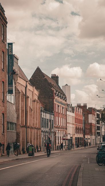 A street scene depicting a row of multi-story residential and commercial buildings in Upper Clapton under a partly cloudy sky. The buildings are constructed with red brick and beige materials, some fe