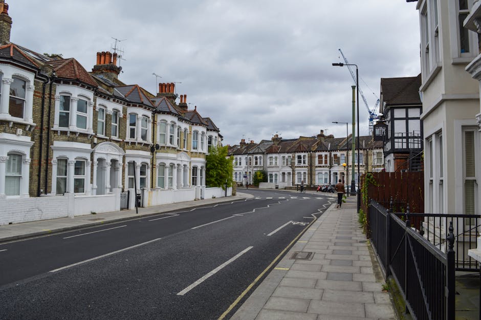 A quiet residential street in Upper Clapton featuring a row of Victorian-style terraced houses with white facades, bay windows, and red-tiled roofs, under a cloudy sky. The pavement runs along the front of the houses, bordered by black metal railings and small front gardens. There is minimal pedestrian activity with a few people walking along the sidewalk. The street has clearly marked parking spaces and a smooth asphalt surface. A streetlamp and a construction crane are visible in the background, indicating ongoing development or maintenance work. The overall scene depicts a typical area suitable for house removals, with no vehicles or furniture in the view, but the setting is relevant to relocation activities carried out by companies like Man with Van Upper Clapton, ensuring optimal logistical planning for furniture transport and packing during home relocations.