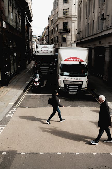 A narrow urban street scene with two white moving trucks parked side by side, one branded with 'uniserve' and the other with 'Becton Transport', positioned on the road with some height clearance, indicating a planned home relocation or furniture transport. A black scooter is parked next to the trucks, and a man in dark clothing and a woman in casual attire are crossing the street in front of the vehicles. The street is flanked by multi-storey buildings with classic architectural facades, and the sky is partly cloudy, with natural daylight illuminating the scene. The image captures the loading process or preparation phase for a house move, consistent with professional removals services by Man with Van Upper Clapton, emphasizing logistics, vehicle positioning, and pedestrian movement within a city environment.